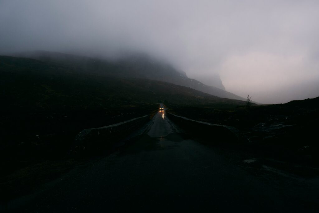 A dull road in a mountainous areas with a singular car driving down it with its headlights on. Mountains can be seen in the background, surrounded by fog and clouds.